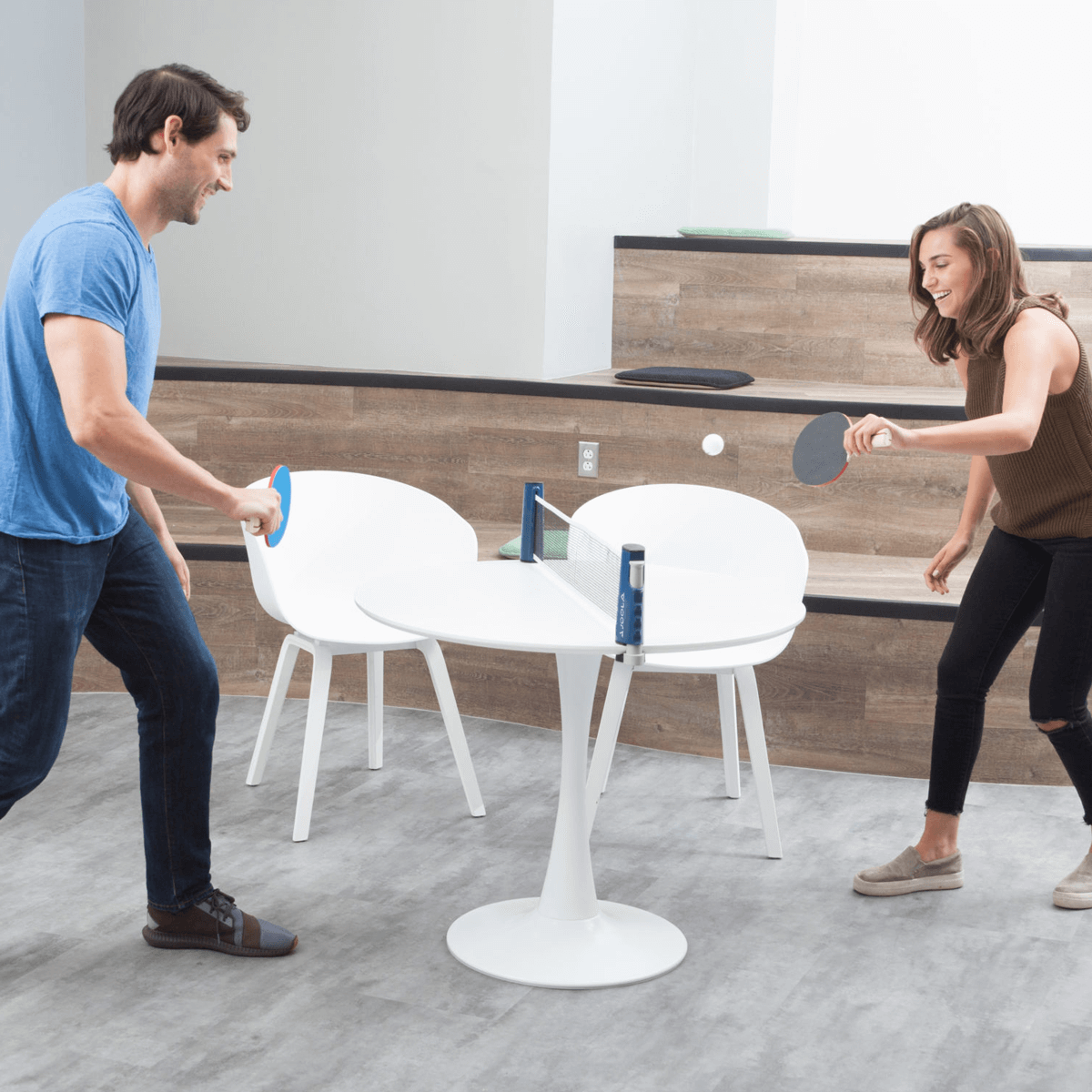 Image: A man and a woman playing table tennis on a small, round cafeteria table in an office setting with the JOOLA Essentials Variant Net Set set up on the table.