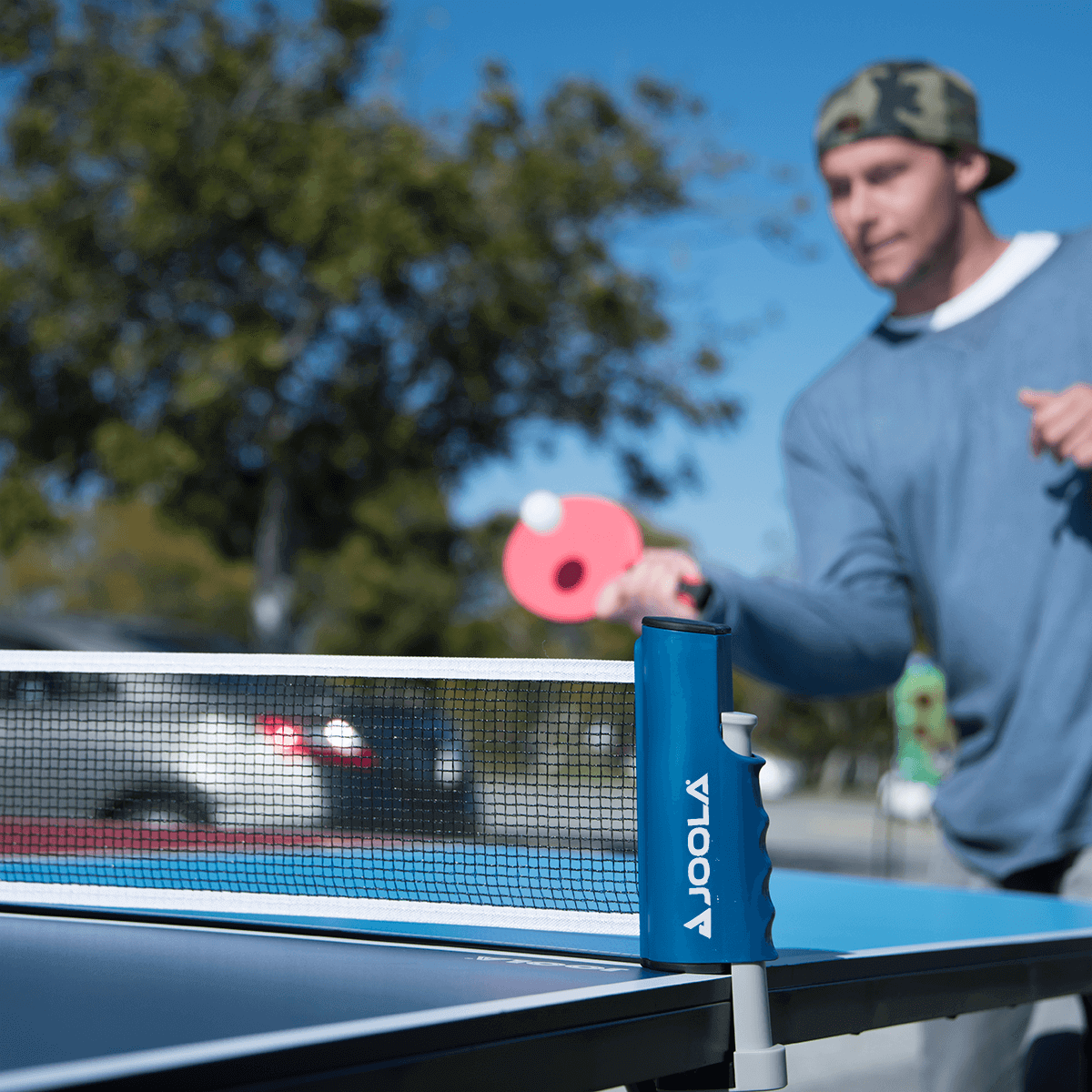 Image: Man playing table tennis in a parking lot outdoors using the Essentials Variant Set. The retractable net is attached to a light blue mini table tennis table.
