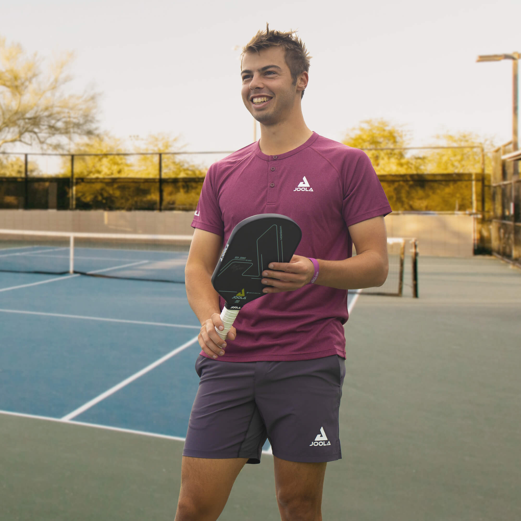 Image: Ben Johns smiling on the pickleball court dressed in the JOOLA Ben Johns Propel Shirt in Burgundy and JOOLA Ben Johns Fluid Shorts in Black