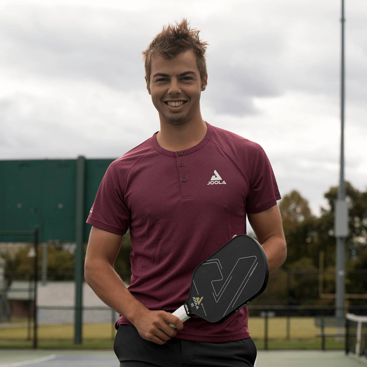 Image: Ben Johns smiling while holding a JOOLA Hyperion pickleball paddle on the court and wearing the JOOLA Ben Johns Short Sleeve Henley in Burgundy